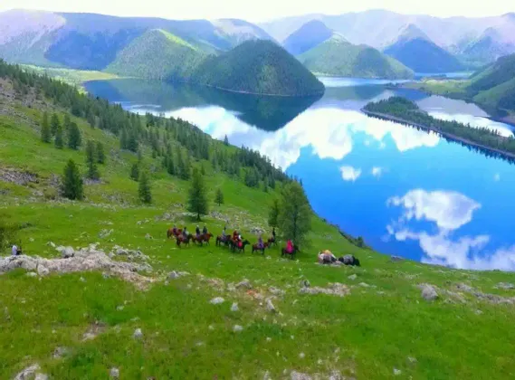 Eine WeRoad Gruppenreise zu Pferd reitet entlang eines grasbewachsenen Hügels, der einen ruhigen See überblickt, in dem sich Himmel und Berge spiegeln.