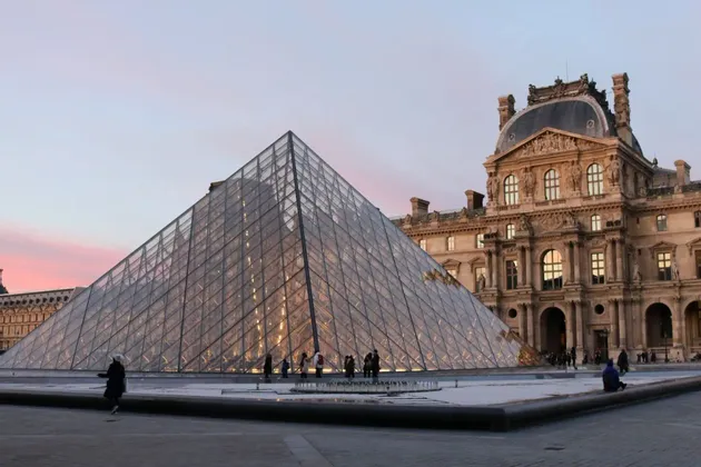 A large glass pyramid stands in a paved courtyard in front of an ornate, historic building under a pink and blue sky at dusk.