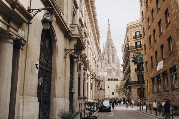 A narrow city street lined with historic stone buildings, with the spire of a Gothic cathedral visible in the distance.