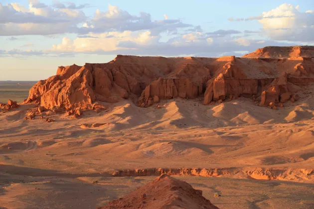 Des formations rocheuses rouges et des collines sablonneuses dans un vaste paysage désertique sont éclairées par le soleil de l'heure dorée sous un ciel partiellement nuageux.