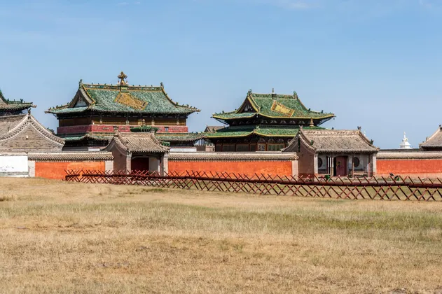 Un monastère mongol aux toits de tuiles vertes et au mur d'enceinte rouge, vu d'un champ d'herbe sèche sous un ciel bleu clair.