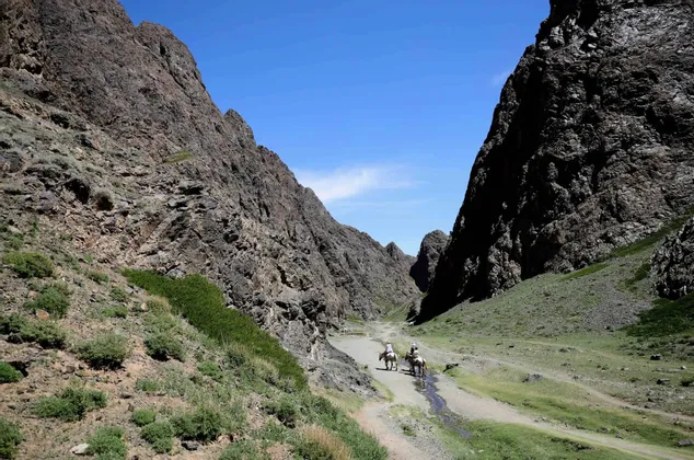 Deux personnes en voyage de groupe WeRoad montent à cheval à travers une vaste vallée montagneuse et rocheuse, près d'un ruisseau sous un ciel bleu.