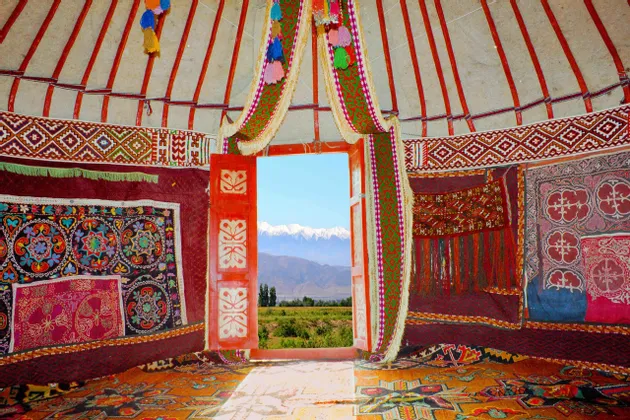 L'intérieur d'une yourte traditionnelle et ornée, avec une porte rouge ouverte encadrant une vue sur des montagnes enneigées.