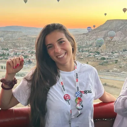 Una donna sorridente con una maglietta WeRoad posa in mongolfiera all'alba, con altre mongolfiere che fluttuano su un paesaggio roccioso.