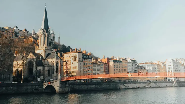 Un vivace ponte pedonale arancione attraversa un fiume di fronte a una città collinare con una grande chiesa ed edifici storici.