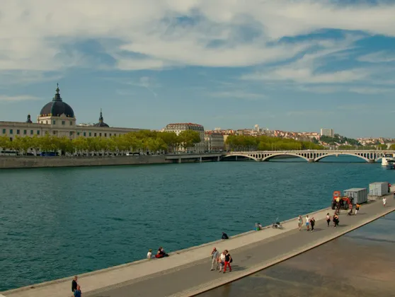 La gente passeggia e si rilassa su un lungofiume in una città, con un edificio a cupola e un grande ponte sullo sfondo.