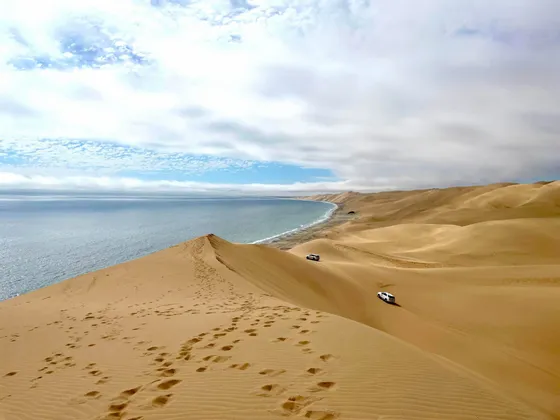 Vaste dune di sabbia digradano verso l'oceano, con due veicoli 4x4 bianchi che percorrono la linea costiera.
