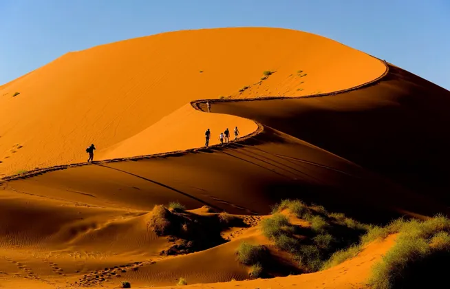 Un gruppo WeRoad in viaggio cammina in fila lungo la cresta di una vasta duna di sabbia arancione sotto un cielo azzurro e limpido.