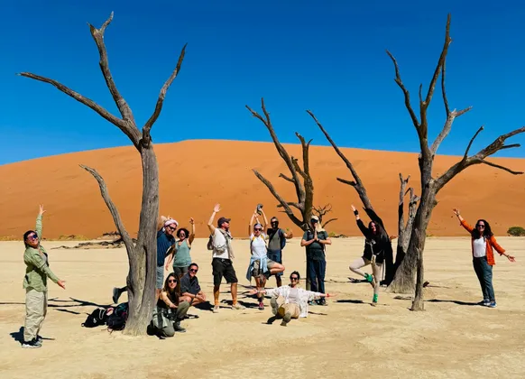 Un viaggio di gruppo WeRoad in posa tra alberi spogli, in un deserto con grandi dune di sabbia arancioni sotto un cielo azzurro.