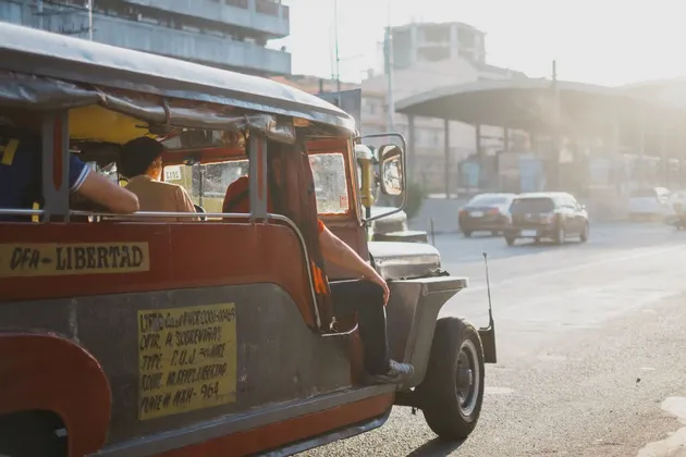 Ein rot-silberner Jeepney transportiert Fahrgäste entlang einer sonnigen Stadtstraße, im Hintergrund Gebäude und andere Autos.
