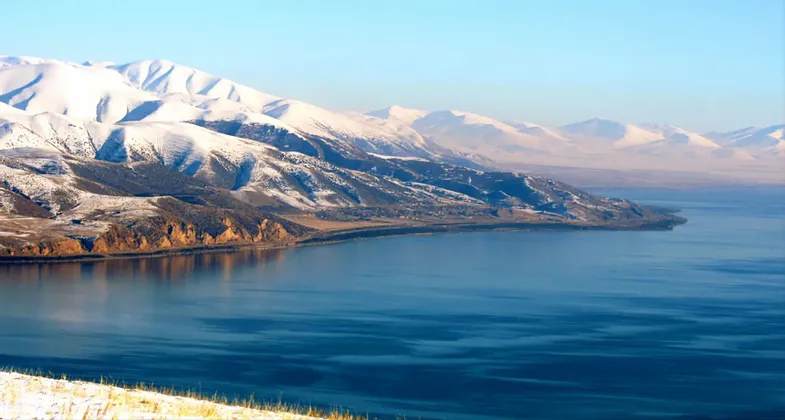 Schneebedeckte Berge treffen am Ufer eines ruhigen, tiefblauen Sees unter klarem Himmel.