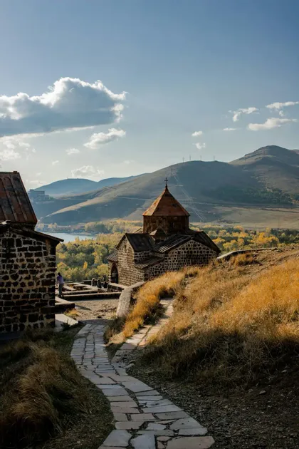 Ein Steinpfad schlängelt sich einen grasbewachsenen Hügel hinauf zu einer alten Steinkirche, die einen See und Berge im Hintergrund überblickt.