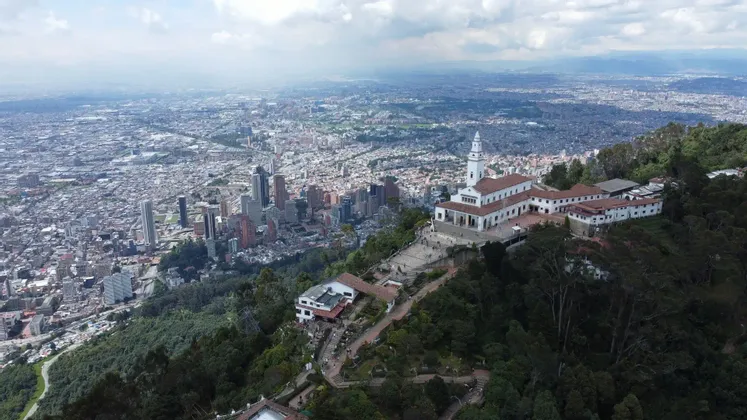 Vista aerea di un santuario bianco su una montagna boscosa che domina una vasta città moderna sotto un cielo nuvoloso.
