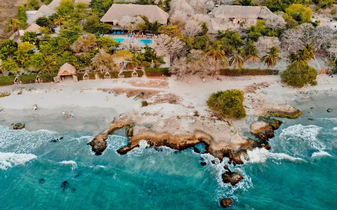 Vista aerea di una costa tropicale dove l'acqua turchese incontra una spiaggia sabbiosa e un resort con edifici dal tetto di paglia tra gli alberi.