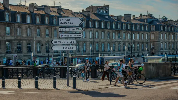 Segnali stradali direzionali bianchi in francese si trovano di fronte a un grande edificio classico, mentre i pedoni attraversano la strada in una giornata di sole.