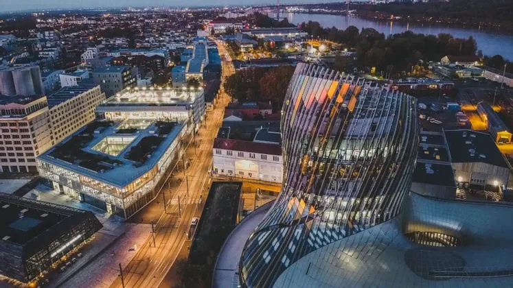 Una vista aerea di un moderno edificio in vetro curvo che si affaccia su una città con strade illuminate e un fiume al crepuscolo.