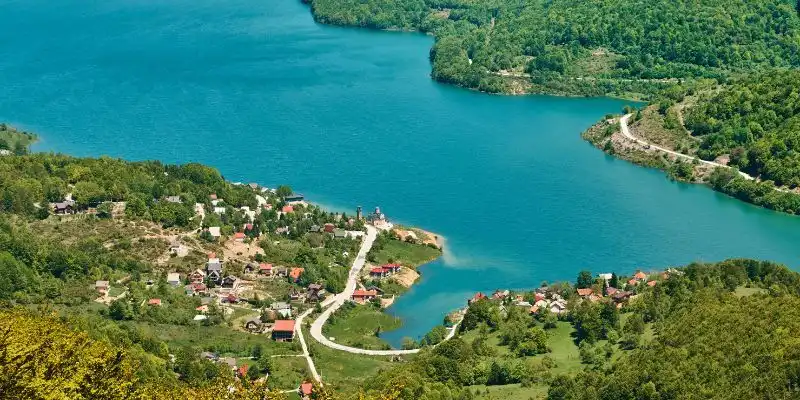 Eine Luftaufnahme eines kleinen Dorfes am Ufer eines hellblauen Sees, eingebettet in dichte, grüne, bewaldete Hügel.