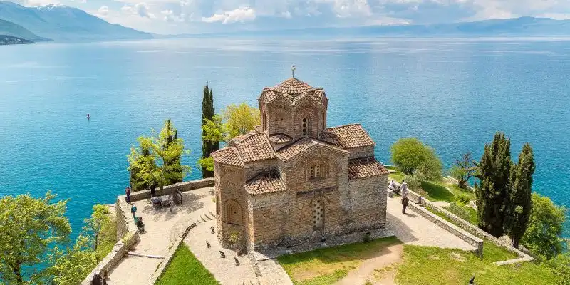 Eine alte Steinkirche mit Ziegeldach thront auf einer grünen Klippe über einem weiten blauen See und fernen Bergen.