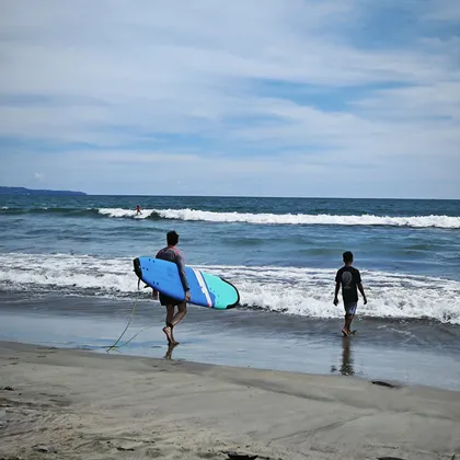 Eine Person mit blauem Surfbrett und ein Junge gehen am Strand entlang in die Meereswellen unter einem teilweise bewölkten Himmel.