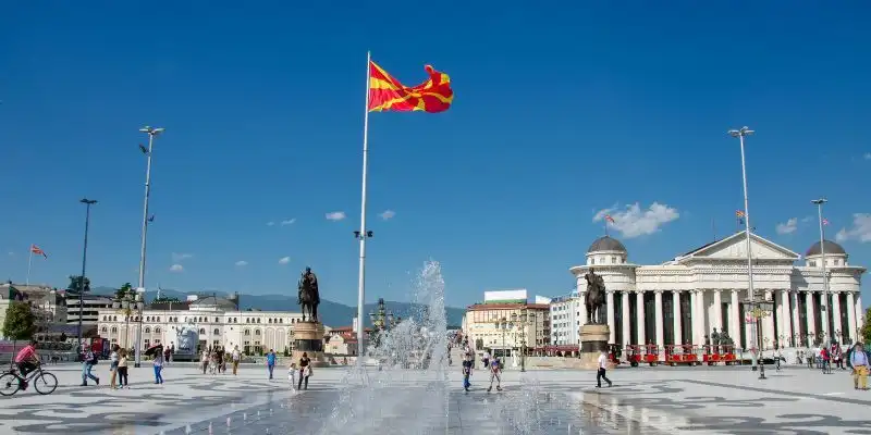 Die rot-gelbe Flagge Nordmazedoniens weht an einem hohen Fahnenmast auf einem belebten Stadtplatz mit Brunnen und Statuen unter einem klaren blauen Himmel.