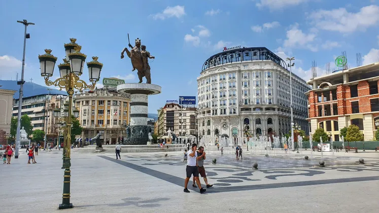 Ein weitläufiger Stadtplatz mit einer zentralen Reiterstatue, einem ebenerdigen Brunnen und Menschen, die unter blauem Himmel zwischen imposanten Gebäuden flanieren.