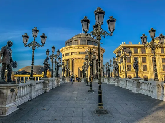 Eine breite Steinbrücke, gesäumt von zahlreichen Bronzestatuen und verzierten Laternen, führt zu einem prächtigen klassischen Gebäude unter blauem Himmel.