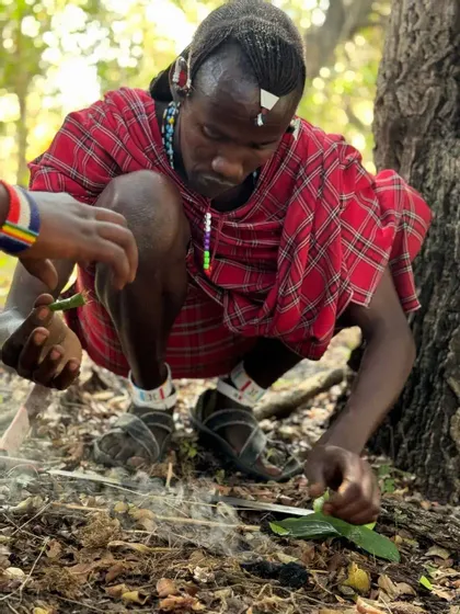 Un uomo con abiti tradizionali Maasai rossi è accovacciato sul suolo della foresta, intento a curare un piccolo fuoco fumante con foglie.