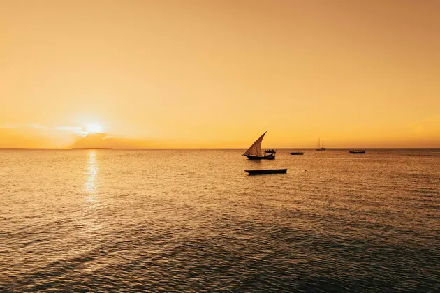 Un tradizionale veliero dhow e altre barche sull'oceano durante un tramonto dorato, con il sole che si riflette sull'acqua.