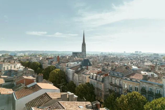 Una vista aerea di una città europea con tetti in terracotta e un'alta guglia di chiesa che si erge al centro sotto un cielo azzurro velato.