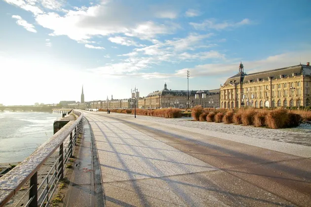 Un ampio lungofiume spolverato di neve si snoda tra edifici storici sotto un cielo azzurro e brillante.