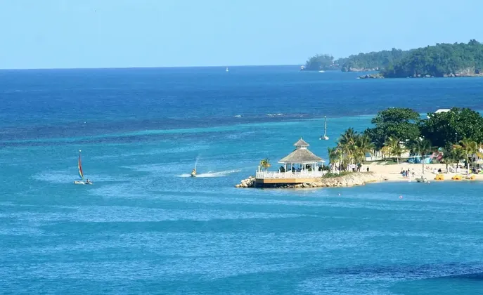 Un kiosque blanc sur une presqu'île domine une plage tropicale avec palmiers et bateaux sur la mer turquoise sous un ciel dégagé.