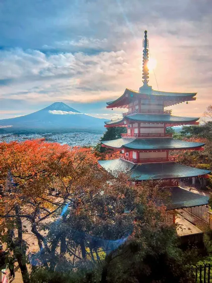 Una pagoda rossa giapponese sorge tra alberi dal fogliame autunnale, affacciata su una città con un grande vulcano sullo sfondo all'alba.
