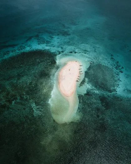 Vista aerea di una piccola isola di sabbia rosa dove sono ormeggiate diverse imbarcazioni, circondata da acqua turchese e una barriera corallina scura.