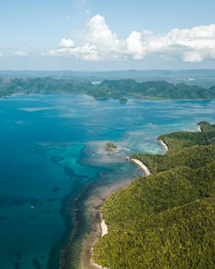 Vista aerea di una costa tropicale con lussureggianti colline verdi che si affacciano su un oceano turchese e isole in lontananza.