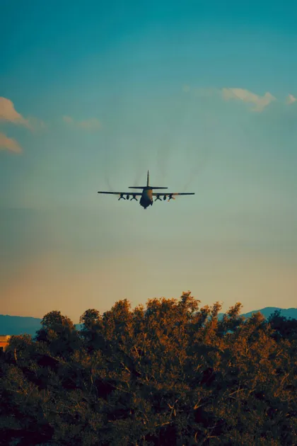 Aereo da trasporto militare visto da dietro in volo a bassa quota sopra le cime degli alberi al tramonto.