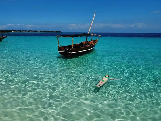 Une femme en bikini vert flotte sur le dos dans une eau turquoise claire à côté d'un bateau en bois traditionnel sous un ciel bleu.