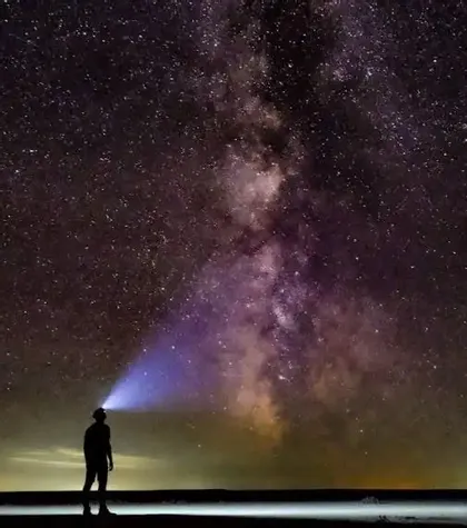 Una persona in silhouette sulla riva illumina la Via Lattea e il cielo stellato con una lampada frontale brillante.