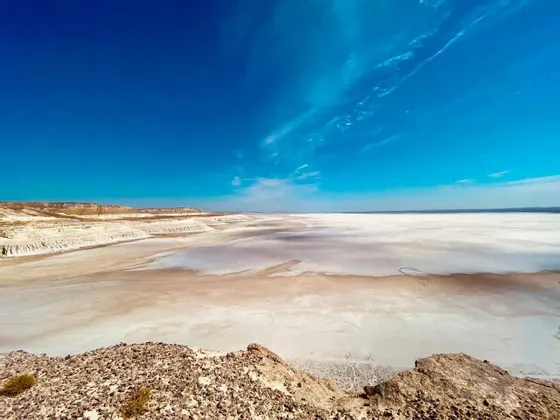 Una vista panoramica da uno sperone di roccia su una vasta e arida distesa salina che si estende all'orizzonte sotto un cielo azzurro intenso.