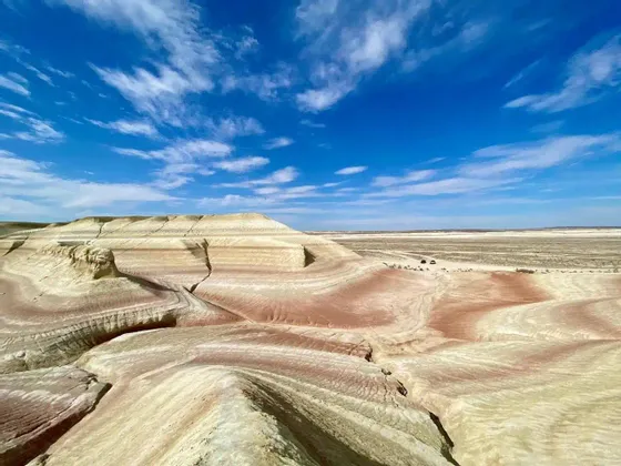 Formazioni rocciose multicolori e stratificate in un vasto paesaggio desertico sotto un cielo azzurro brillante con nuvole, con due veicoli in lontananza.