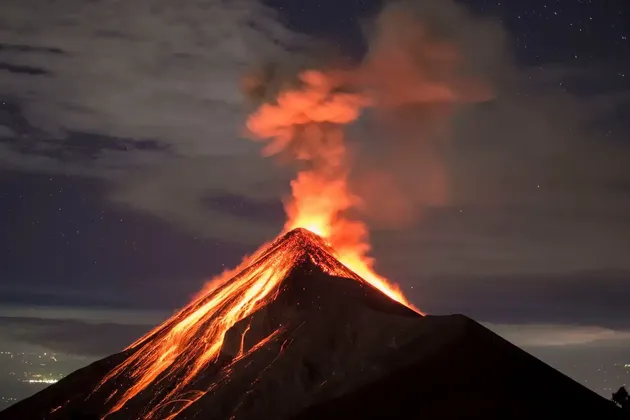 A volcano erupts at night, spewing glowing lava and smoke under a starry sky with clouds.