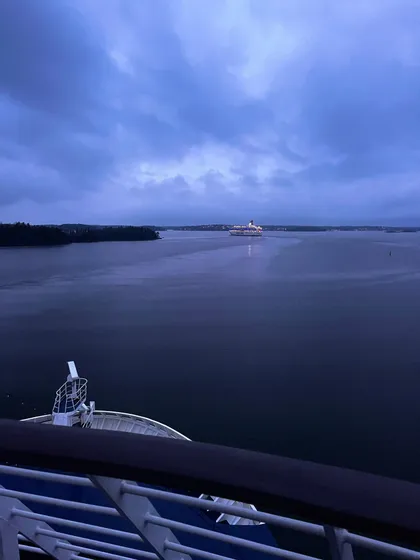 Dal ponte di una nave, si ammira un'altra grande imbarcazione navigare sull'acqua al tramonto, con una costa boscosa sullo sfondo.