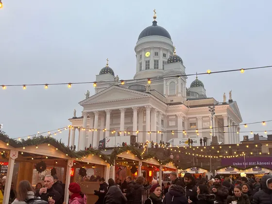 Un vivace mercatino di Natale con bancarelle in legno e luminarie, con una grande cattedrale bianca sullo sfondo sotto un cielo nuvoloso.