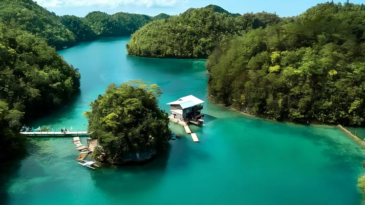 Una vista aerea di una casa moderna su un piccolo isolotto in un fiume turchese, circondata da colline lussureggianti e alberate.