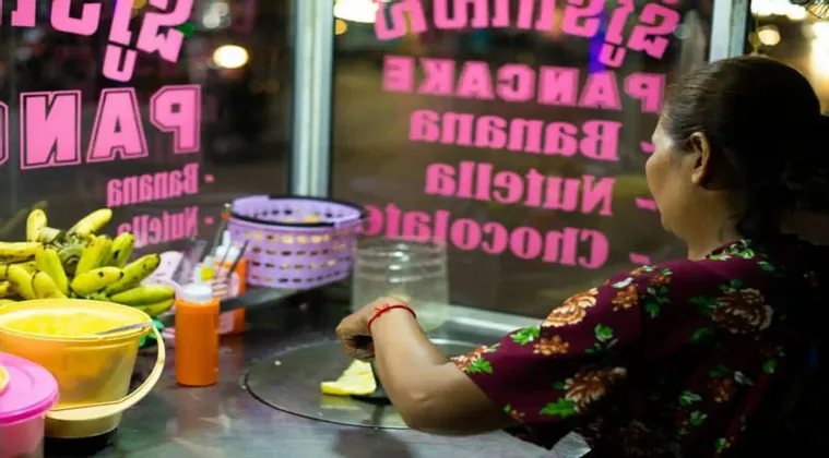 Una mujer en un puesto de comida callejera por la noche hace un panqueque en una plancha junto a un racimo de plátanos.