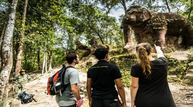 Una mujer de un viaje en grupo de WeRoad señala una gran estatua de elefante de piedra cubierta de musgo mientras dos hombres observan en un bosque.
