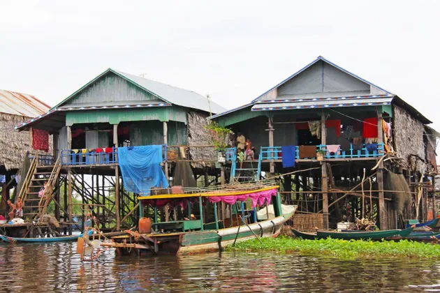 Casas de madera sobre pilotes de un pueblo flotante se alzan sobre el agua, con barcos amarrados y personas visibles en los porches.