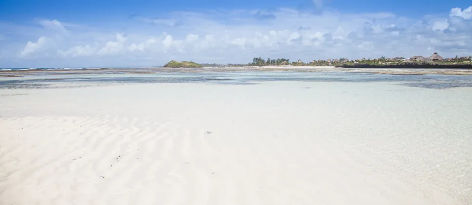 Una vasta distesa di sabbia bianca con increspature è visibile sotto acqua bassa e limpida, con una costa tropicale in lontananza sotto un cielo azzurro.