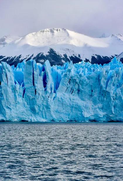 Un enorme glaciar azul se alza sobre las aguas oscuras, con montañas cubiertas de nieve al fondo.