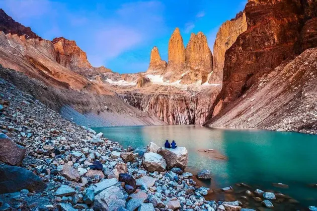 Due persone siedono su un grande masso sulla riva di un lago turchese, ai piedi di drammatiche guglie montane illuminate dal sole.