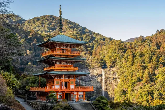 Una pagoda giapponese arancione a tre piani si erge su un fianco della montagna, accanto a un'alta cascata, circondata da una fitta vegetazione autunnale.